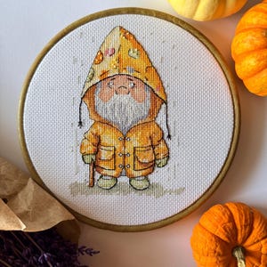 May include: A cross-stitch embroidery of a gnome wearing a yellow hooded coat, holding a walking stick. The embroidery is set in a round wooden hoop. The image is surrounded by small pumpkins and lavender.
