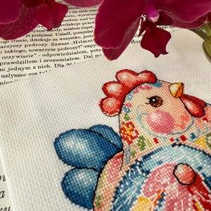 May include: A colorful cross-stitch rooster design with blue, pink, and yellow accents. The rooster has a red comb and wattle, and is set against a white background. The image is partially framed by a dark red orchid.