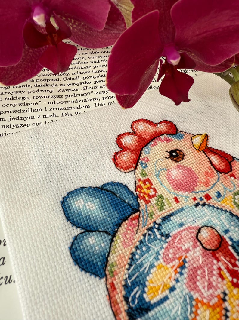 May include: A colorful cross-stitch rooster design with blue, pink, and yellow accents. The rooster has a red comb and wattle, and is set against a white background. The image is partially framed by a dark red orchid.