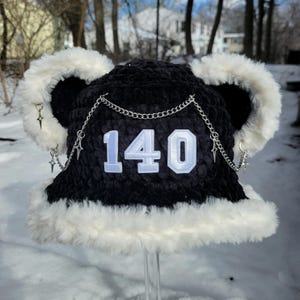 May include: A black crochet hat with white faux fur trim and ear accents. The hat features the number "140" in light blue and white, with a silver chain and star charms. The hat is designed for cold weather.