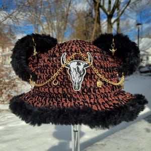 May include: A brown and black patterned bucket hat with black faux fur trim and ear accents. The hat features a white bull skull patch, gold chain, and dangling charms. The hat is displayed outdoors in a snowy environment.