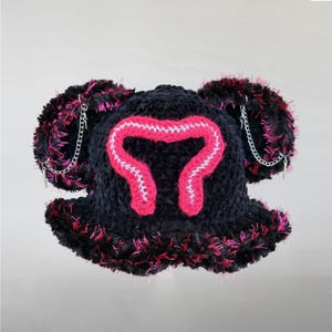 May include: Black crochet hat with a pink and white design on the front. The hat has fluffy black and pink trim and ear-like accents with silver chain details. A unique, handmade accessory.
