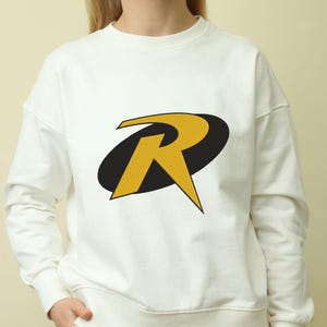 May include: White sweatshirt featuring a large yellow "R" logo with a black oval background. The logo is centered on the front of the long-sleeved, crew neck top. The sweatshirt is made of a soft material.