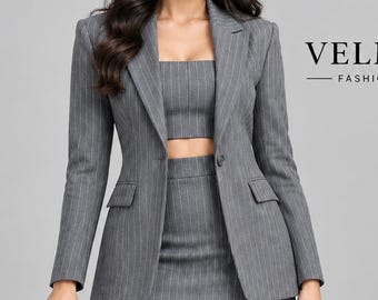 May include: A grey pinstriped blazer, a matching cropped top, and a high-waisted mini skirt. The set features a single-button closure on the blazer and two flap pockets. The text VELMORA FASHION STUDIO is visible in the upper right corner.