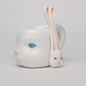 May include: A white ceramic mug shaped like a human face with a blue eye and pink accents. A rabbit-shaped spoon with pink-lined ears and blue eyes rests against the mug. The mug has a handle.