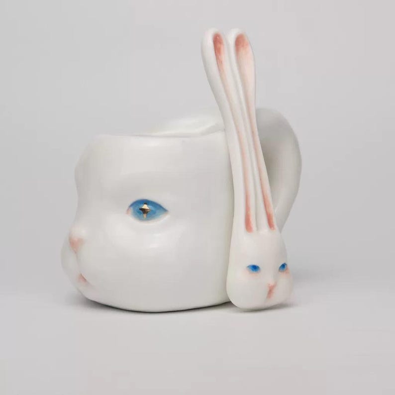 May include: A white ceramic mug shaped like a human face with a blue eye and pink accents. A rabbit-shaped spoon with pink-lined ears and blue eyes rests against the mug. The mug has a handle.