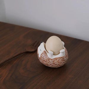 以下が含まれることがあります： ベージュの卵が入った、茶色と白のデザインのセラミックハリネズミの卵カップ。 ハリネズミは笑顔で仰向けになっています。 木製のフォークが背景にあります。 卵カップは濃い木製の表面にあります。