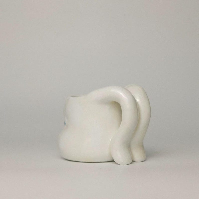 May include: A white ceramic mug with a unique design. The mug features a rounded body and two curved, elongated handles. A small, light blue detail is visible on the side of the mug. The mug is set against a plain white background.