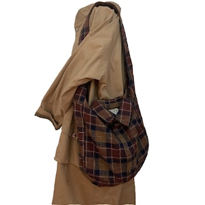 May include: A shoulder bag with a plaid pattern in burgundy, navy, and gold hangs on a tan coat. The bag is made from a button-down shirt, with a collar and button closure. The strap is made from the same plaid fabric.