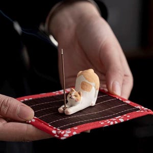 May include: A ceramic cat incense holder in white and orange, with a brown incense stick. The cat is lying on a rectangular bamboo mat with a red border. The holder is held in a person's hands.
