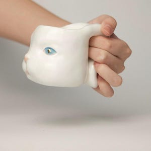 May include: A white ceramic mug shaped like a cat's head, held in a hand. The mug features blue eyes and a pink nose. The handle is designed as the cat's ear. The mug is a unique and whimsical item.