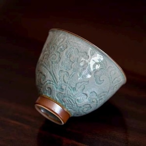 Puede incluir: Una taza de té de cerámica azul claro con un patrón floral en relieve en su superficie exterior. El esmalte muestra una textura fina de grietas, mientras que el borde y la base circular tienen un acabado en un tono marrón liso y sólido.
