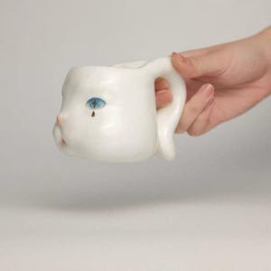 May include: A white ceramic mug with a face design. The mug features a blue eye with a teardrop and a pink nose. The handle is shaped like an ear. The mug is held by a hand.