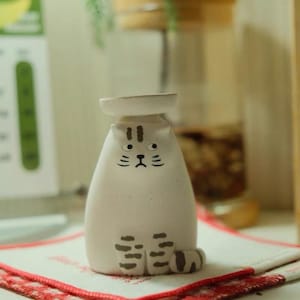 Könnte beinhalten: Eine weiße Keramik-Katzenfigur mit minimalistischem Design. Die Katze hat schwarze Gesichtszüge und gestreifte Pfoten. Sie sitzt auf einem rot-weiß karierten Tuch. Auf ihrem Kopf befindet sich ein kleiner, runder, weißer Teller.