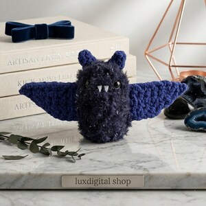 Puede incluir: Un murciélago de peluche azul oscuro con alas extendidas y colmillos blancos. El murciélago está hecho de un material texturizado y moteado. Un lazo azul oscuro y libros están en el fondo. Un objeto decorativo.