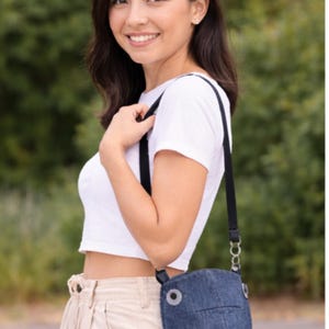 May include: A denim shark-shaped crossbody bag with a black strap. The bag has a round eye and fin details. The person is wearing a white crop top and khaki pants.