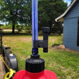 May include: A red gas can with a black cap and a blue tube attached. A black nozzle is connected to the tube. The can is outdoors with a green lawn and trees in the background.