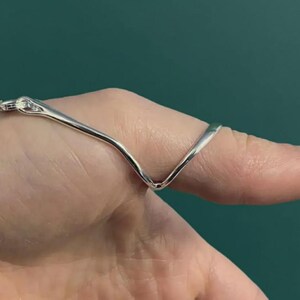 Puede incluir: Un anillo plateado con un dise&ntilde;o en forma de V, conectado a una pulsera de cadena. El anillo se lleva en un dedo, sobre un fondo verde azulado.