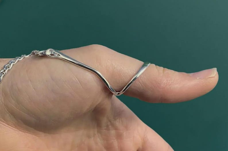 Puede incluir: Un anillo plateado con un dise&ntilde;o en forma de V, conectado a una pulsera de cadena. El anillo se lleva en un dedo, sobre un fondo verde azulado.