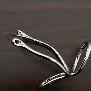 Puede incluir: Una escultura de metal plateado pulido con un dise&ntilde;o abstracto y fluido. La pieza presenta l&iacute;neas curvas y bucles, creando una sensaci&oacute;n de movimiento. La escultura est&aacute; sobre un fondo de madera oscura.