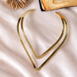 May include: A gold-coloured, hammered metal collar necklace. The necklace has a V-shape design with two parallel lines. The metal has a textured surface and a shiny finish. The necklace is displayed on a soft, pink surface.