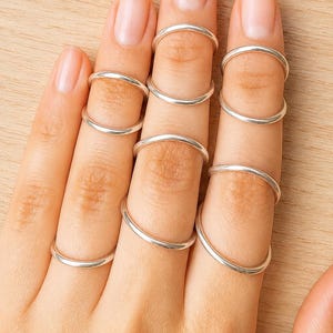 May include: A set of seven thin, smooth, polished silver-tone metal rings worn on the fingers. Each ring features a simple, minimalist circular band design with a reflective metallic finish, positioned across the fingers in a stacked arrangement.