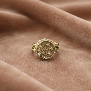 Könnte beinhalten: Ein goldfarbener Ring mit einem Himmelsdesign. Der Ring hat eine runde Oberfläche mit einem Mond und Sternen sowie aufwendigen Details. Der Ring ist vor einem weichen, rosabraunen Hintergrund platziert.