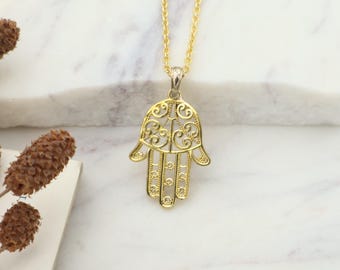 May include: Gold-coloured Hamsa hand pendant necklace. The pendant has an openwork design with detailed patterns. The necklace features a fine chain and a lobster clasp. The Hamsa hand is a symbol of protection and good luck.