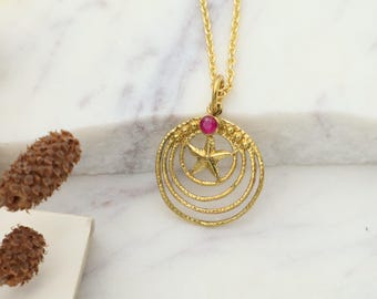 May include: Gold-toned pendant necklace featuring a circular design with a star in the center. The pendant has a small, round, red gemstone at the top. The necklace hangs on a delicate gold chain, creating an elegant and eye-catching accessory.
