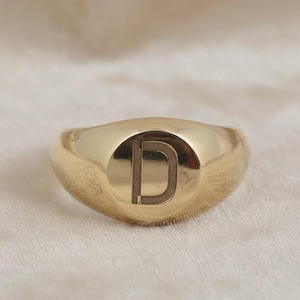 Op de afbeelding: Een goudkleurige zegelring met de letter "D" gegraveerd op een cirkelvormig vlak. De ring heeft een gladde, gepolijste afwerking en een afgeronde band. De achtergrond is een zachte, crèmekleurige stof.