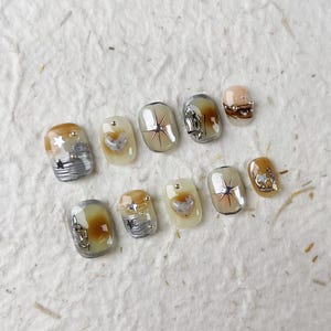 Peut inclure: Un ensemble de dix faux ongles avec divers motifs. Les ongles présentent un mélange de couleurs, dont l'argent, l'or et le beige. Certains ongles ont des motifs d'étoiles et de cœurs, tandis que d'autres ont un effet marbré. Les ongles sont disposés sur une surface texturée blanc cassé.