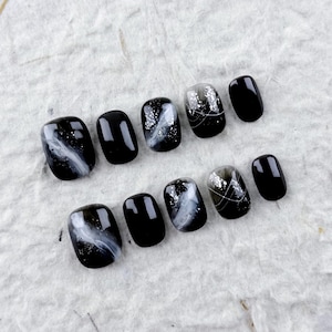 May include: A set of ten black and white press-on nails. The nails feature a glossy finish with designs including a white marble effect, silver glitter, and silver line accents. The nails are arranged on a textured white surface.