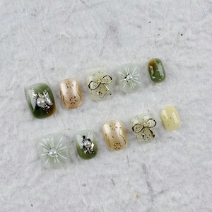 Peut inclure: Un ensemble de dix faux ongles aux designs variés. Les ongles présentent un mélange de couleurs vertes, dorées et transparentes, ornés de nœuds, de perles et de motifs floraux. Ils sont disposés sur une surface texturée blanc cassé, pour la manucure.