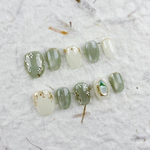 以下が含まれることがあります： 緑と白の様々な色合いのつけ爪のセット。一部の爪には金箔のアクセントと繊細な白い花の模様があります。爪は、質感のあるオフホワイトの表面に配置されています。