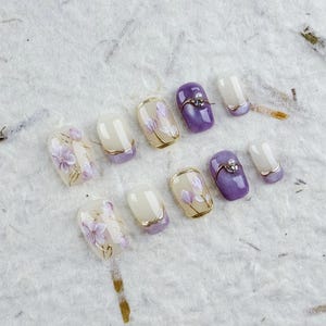 Pode incluir: Um conjunto de unhas postiças com um design floral. As unhas apresentam uma variedade de cores, incluindo branco, roxo e dourado. Algumas unhas têm uma borda dourada, outras prateada. As unhas estão dispostas sobre uma superfície branca texturizada.