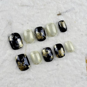Peut inclure: Un ensemble de faux ongles avec des motifs noirs et crème. Certains ongles présentent des motifs floraux dorés, d'autres sont unis. Les ongles sont présentés sur une surface texturée blanc cassé.
