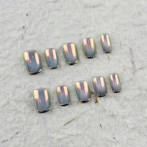 May include: A collection of iridescent false nails. The nails are rectangular with rounded edges and a delicate gold trim. The colour shifts between blue, pink, and yellow, creating a shimmering effect. The nails are arranged in two rows.