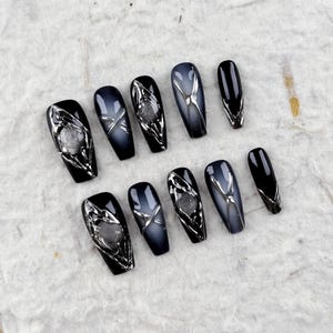 May include: A set of coffin-shaped press-on nails. The nails feature a gradient design, transitioning from black to a smoky gray. Some nails have silver metallic embellishments, while others are solid black. The nails are arranged on a textured white surface.
