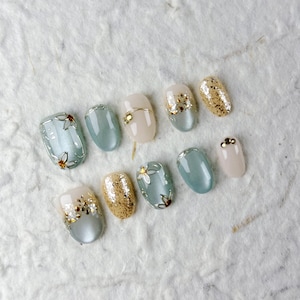 Puede incluir: Una colección de uñas artificiales de forma ovalada con varios diseños. Algunas son de color azul claro, otras con purpurina dorada y otras de color beige pálido. Varias uñas presentan detalles florales dorados y purpurina dorada. Las uñas están dispuestas sobre una superficie blanca texturizada.