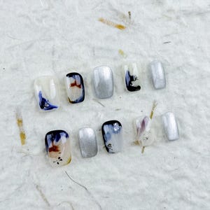 Puede incluir: Un conjunto de diez uñas postizas de forma cuadrada. Los diseños incluyen purpurina plateada, patrones abstractos de acuarela en azul, marrón y negro, y pequeños detalles florales. Las uñas están dispuestas sobre una superficie texturizada de color blanco roto.