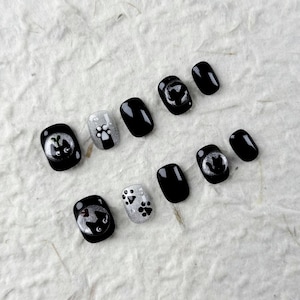 May include: A collection of black and silver press-on nails. Some nails display a cat face design, others feature paw prints. The nails are oval-shaped with a glossy finish. The background is a textured, off-white surface.