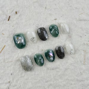 May include: A collection of press-on nails in various designs. The nails feature colors like silver, dark green, white, and black. Some nails have glitter, pearl accents, or textured silver foil. The nails are arranged on a textured, off-white surface.