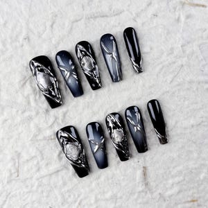 Puede incluir: Un conjunto de diez uñas postizas en forma de ataúd. Las uñas son negras, grises y plateadas. Algunas uñas tienen adornos plateados, mientras que otras tienen un efecto degradado. Las uñas están dispuestas sobre una superficie blanca texturizada.