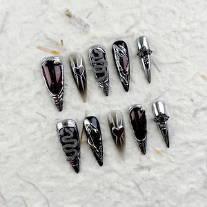 Puede incluir: Un conjunto de diez uñas postizas largas y puntiagudas. Las uñas presentan una variedad de diseños, incluyendo detalles de serpientes y corazones plateados, y esquemas de color rojo oscuro y negro. Algunas uñas tienen un acabado plateado metálico.
