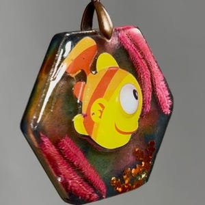 May include: A hexagonal pendant featuring a cartoon fish with yellow and orange stripes. The pendant is encased in clear resin with pink and orange accents. A bronze-colored bail is attached at the top.