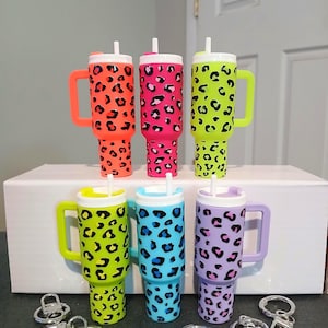 May include: Six colorful tumblers with handles and straws, featuring a leopard print design. The tumblers come in neon orange, pink, and green, with additional options in blue and purple. Silver keychains are in the foreground.