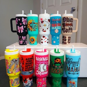 May include: A collection of insulated tumblers with various designs and colors. Some tumblers feature text like "Mama needs coffee" and "Blessed Mama." Other designs include sunflowers, bows, and baseball themes. Each tumbler has a handle or a straw.
