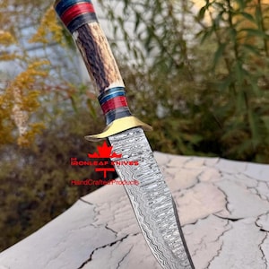 Peut inclure: Un couteau à lame fixe avec une lame en acier Damas au motif ondulé. Le manche est en bois de cerf texturé avec des entretoises rouges, bleues et noires et une garde en laiton. La lame porte une feuille d'érable rouge et IRONLEAF KNIVES.