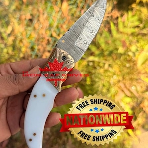 May include: A folding knife with a Damascus steel blade featuring a wavy pattern, a brass bolster with an engraved floral design, and a smooth white handle with three small brass pins. The blade is marked with the text IRON OF KNIVES.