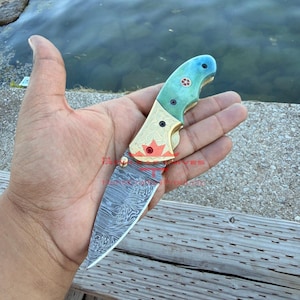 May include: A folding pocket knife with a Damascus steel blade, a brass bolster featuring an engraved maple leaf, and a teal-coloured handle. The knife includes a small gold-coloured thumb stud and a decorative circular inlay on the handle grip.
